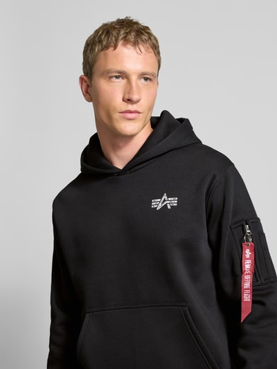 Männermodel trägt Hoodie mit Label-Details von Alpha Industries in Schwarz, Baumwoll-Mix, lockerer Schnitt, Nahaufnahme Oberkörper.