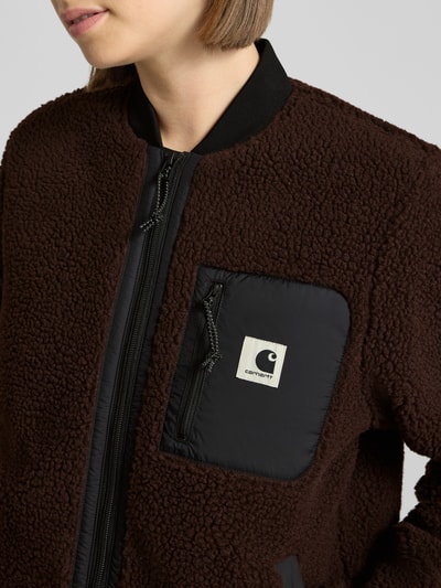Frauenmodel trägt Fleecejacke mit Zweiwege-Reißverschluss Modell 'Janet' von Carhartt Work In Progress in Braun mit Teddyfell und Kontrast-Brusttasche, Nahaufnahme Oberkörper. Damen Fleecejacke, Regular Fit, Stehkragen, Label-Detail, Eingrifftaschen, Kontext: Rückenlänge 80 cm, Schulterbreite 38 cm, Brustweite 52 cm, Ärmellänge 62 cm.