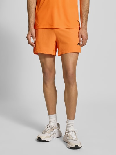 Männermodel trägt Regular Fit Shorts mit Logo-Print Modell 'Power' von adidas Training in Orange mit Strukturmuster, Eingrifftaschen, Nahaufnahme Oberschenkel vorne.