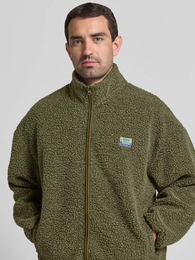 Männermodel trägt groene fleecejack met opstaande kraag en labeldetail model 'HOKTOWN' van American Vintage, relaxed fit, ritssluiting, steekzakken, ruglengte maat M/L 69 cm, schouderbreedte 56 cm, borstwijdte 48 cm, mouwlengte 55 cm, textuur met grove lussen, nahaufnahme bovenlichaam.