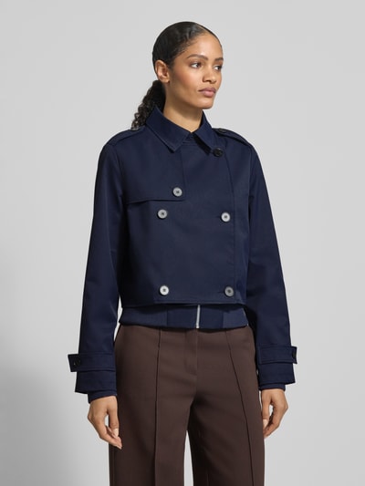 Vrouwenmodel draagt Trenchcoat mit Ärmelriegeln van monari in donkerblauw, regular fit, dubbele rij knopen, kraag, knoopsluiting, details op mouwen, voorkant volledig zichtbaar, bovenlichaam close-up.