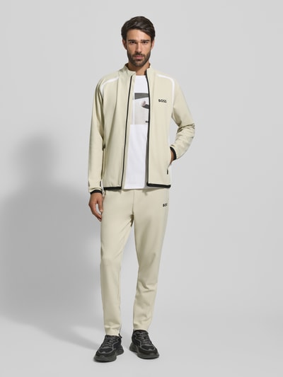 Männermodel trägt Regular Fit Trainingsjacke mit Passeinsätzen Modell 'TOC SPIN FZ' von BOSS Green in Optik: Unifarben mit Label-Print, Stehkragen, Langarm und seitlichen Reißverschlusstaschen, Materialverarbeitung: Twill, Ganzkörperansicht von vorne.