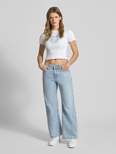 Modelka nosi T-Shirt mit Label-Print von Levi's®, biała bawełna, krój slim fit, okrągły ściągacz, falbanki, nadruk, marszczenia, pełny widok z przodu.