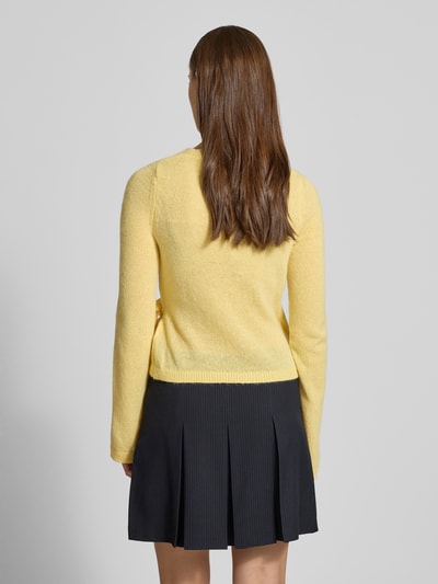 Vrouwenmodel draagt Strick-Cardigan in wikkellook van Review, regular fit, V-hals, lange mouwen, sluiting met strik, ruglengte 50 cm, mouwlengte 66 cm, zwarte geplooide rok, effen lichtgeel gebreid materiaal, rugaanzicht.