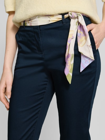 Frauenmodel trägt Slim Fit Chino mit Viskose-Anteil von Christian Berg Woman in Dunkelblau, florales Stoffband, Nahaufnahme Oberkörper. Elastischer Baumwoll-Mix, verkürzte Passform, Knopf- und Reißverschluss sichtbar.