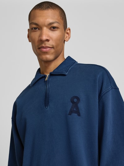 Männermodel trägt Relaxed Fit Troyer aus reiner Bio-Baumwolle Modell 'ICONIC Å GMT' von Armedangels in Uniblau mit Logo-Stitching, Sweat-Textur, Nahaufnahme Oberkörper.