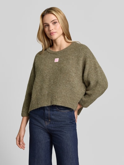 Frauenmodel trägt Oversized Pullover mit Alpaka-Anteil Modell 'East' von American Vintage in Olivgrün, Strick-Optik, Logo-Patch, Nahaufnahme Oberkörper.