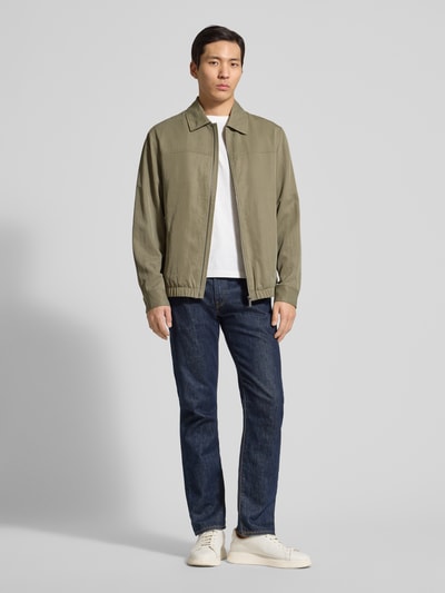 Männermodel trägt Regular Fit Jacke aus Lyocell-Mix von Guess in Olivgrün, sichtbar Umlegekragen und Reißverschlusstaschen, Ganzkörperansicht von vorne.