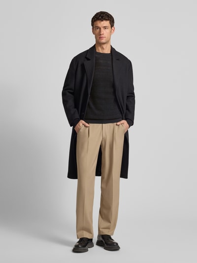 Männermodel trägt Regular Fit Strickpullover mit Rundhalsausschnitt von Antony Morato aus reiner Baumwolle mit Strukturmuster in Schwarz, kombiniert mit cremefarbener Hose und dunklem Mantel, Ganzkörperansicht von vorne.