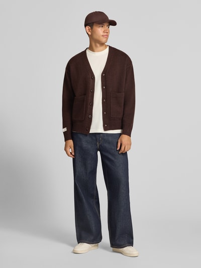 Männermodel trägt Cardigan mit rückseitigem Motiv und V-Ausschnitt von ANOTHER COTTON LAB in Braun, gerade Form, aufgesetzte Taschen, Ganzkörperansicht von vorne.