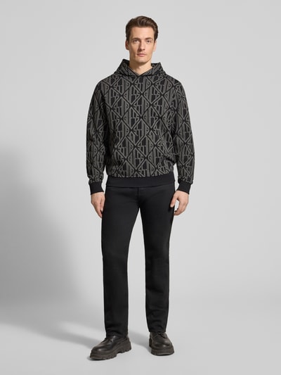 Männermodel trägt Hoodie im Allover-Look mit Känguru-Tasche von Guess aus reiner Baumwolle, Regular-Fit, Ganzkörperansicht von vorne.