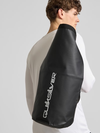 Männermodel trägt Tasche mit Riemen und Label-Print von Quiksilver in Schwarz mit weißem Schriftzug, Nahaufnahme Oberkörper.