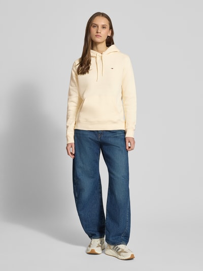 Modelka nosi Regular Fit Hoodie aus Baumwoll-Mix od Tommy Jeans, kolor jednolity, logo, kieszeń kangurka, kaptur ze sznurkiem, krój regular fit, długi rękaw, bawełniany mix, widok całej sylwetki z przodu.
