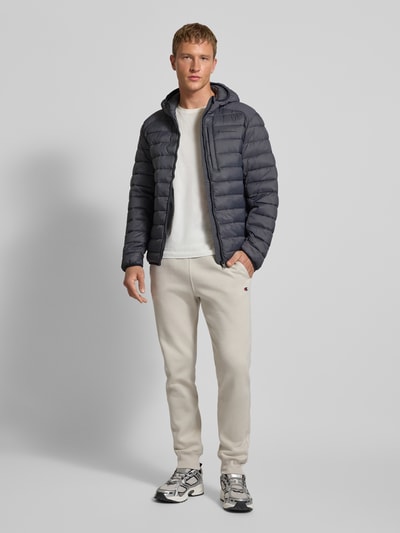Mannenmodel draagt Gewatteerd jack met capuchon en logoprint van CHAMPION, regular fit, ritssluiting, steekzakken, kleur donkergrijs. Ganzkörperansicht von vorne. Ruglengte 71 cm, schouderbreedte 51 cm, borstwijdte 61 cm, mouwlengte 69 cm, maat M.
