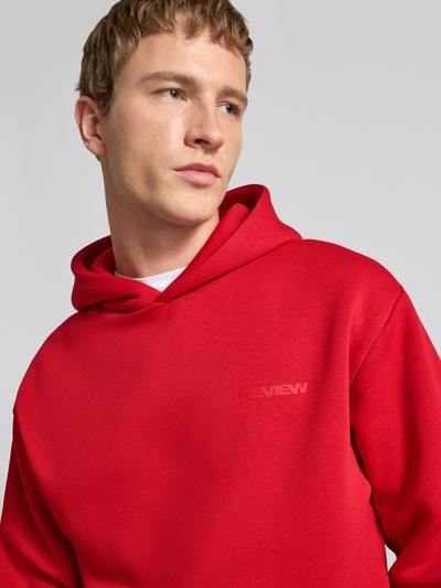 Męski model nosi Regular Fit Hoodie mit Logo-Print od Review, kolor czerwony, bawełna, kaptur, zbliżenie na górną część ciała.