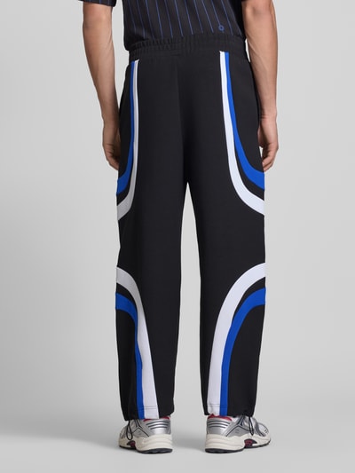 Männermodel trägt Relaxed Fit Sweatpants aus Baumwoll-Mix Modell 'Nalotron' von Hugo Blue in Schwarz mit blauen und weißen Kontraststreifen, elastischer Bund, Sweatmaterial, Ganzkörperansicht Rückansicht.