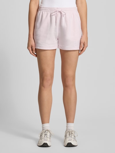 Frauenmodel trägt Straight Fit Shorts mit Tunnelzug Modell 'French' von New Balance aus Baumwoll-Mix in zartem Rosa, Ganzkörperansicht von vorne. Damen Shorts mit elastischem Bund, Bundweite bei Größe S: 40 cm, Innenbeinlänge: 8 cm.