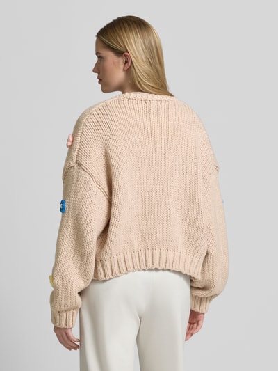 Frauenmodel trägt Oversized Strickjacke mit floralem Zierbesatz von Apricot, Grobstrick, gerippte Abschlüsse, Rückansicht, Ganzkörperansicht. Rückenlänge bei Größe S: 60 cm, Ärmellänge bei Größe S: 51 cm, überschnittene Schultern.