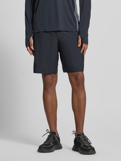 Mężczyzna nosi Tech Utility Shorts mit Label-Print von Under Armour, prosty krój, elastyczny pas, czarny, szerokość w rozmiarze M: 37 cm, długość wewnętrzna nogawki w rozmiarze M: 22 cm, styl utility, nadruk z logo, całkowity widok z przodu.
