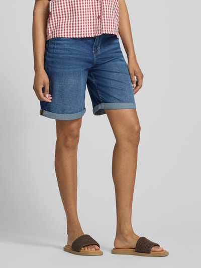 Kobiece modelka nosi Regular Fit Jeansshorts aus Baumwoll-Mix Modell 'SURI' von s.Oliver RED LABEL, denim średni niebieski, krój regular fit, pięć kieszeni, nity, szlufki, podwijane nogawki, detal z metką, zapięcie na guzik i kryty zamek, materiał bawełniany, widok całej sylwetki z przodu.