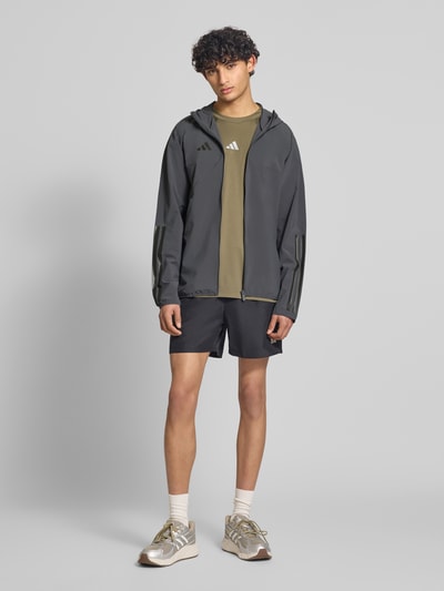 Mężczyznamodel nosi Slim Fit Funktionsjacke aus schnelltrocknendem Material od adidas Sportswear, jednolity kolor, krój slim fit, kaptur, zamek błyskawiczny, nadruk z logo, paski w kontrastowym kolorze, długi rękaw, szybkoschnący, regulujący pot, boczne kieszenie zapinane na zamek błyskawiczny, widok całej sylwetki z przodu.
