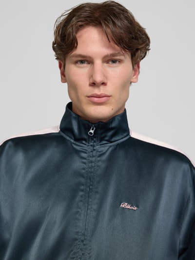 Männermodel trägt Schimmernde Trainingsjacke mit Label-Stitching von BLKVIS in Dunkelblau, Stehkragen, Reißverschluss, Nahaufnahme Oberkörper. Regular-Fit Herren Trainingsjacke, Rückenlänge 61 cm, Ärmellänge 63 cm, glänzende Textur.