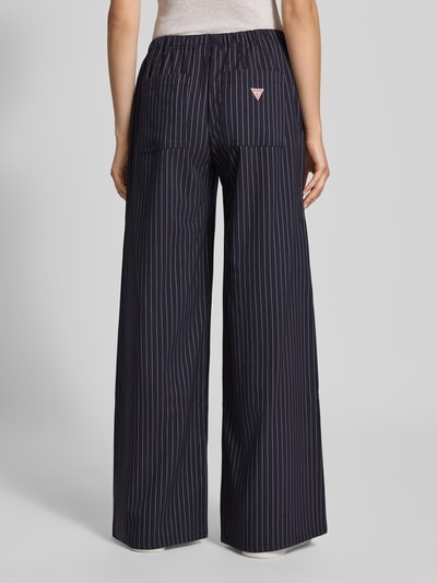 Vrouwenmodel draagt Wide Fit broek met elastische band van GUESS, donkerblauw met witte strepen, bandplooien, bandwijdte 41 cm, binnenbeenlengte 80 cm, stoffen broek met tunnelkoord, bandplooibroek, jeanslook, volledige lichaamsaanzicht achterkant.