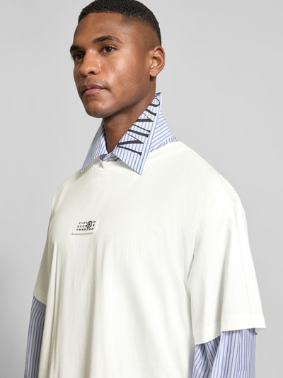 Männermodel trägt Relaxed Fit T-Shirt mit Logo-Detail von MM6 Maison Margiela über gestreiftem Hemd, Weiß mit Rundhalsausschnitt, Nahaufnahme Oberkörper.