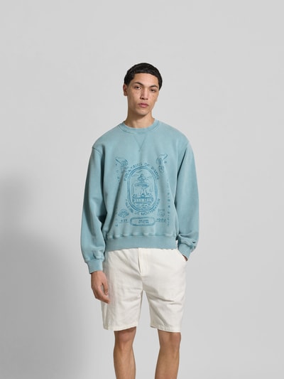 Männermodel trägt Oversized Sweatshirt mit Statement-Print von MAISON KITSUNÉ in Blau im Used-Look, kombiniert mit hellen Shorts, Ganzkörperansicht von vorne.