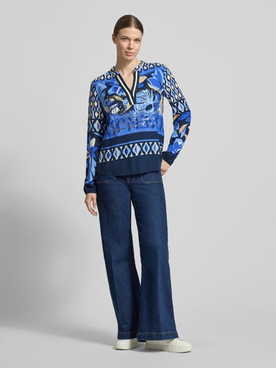 Vrouwenmodel draagt Blusenshirt aus Viskose mit Tunikakragen van Betty Barclay, zuiver viscose, regular fit, all-over look, tuniekkraag, V-hals, ruglengte maat 36: 67 cm, mouwlengte maat 36: 65 cm, blauw met grafisch patroon, wijde jeans, witte sneakers, geheel van voren bekeken.