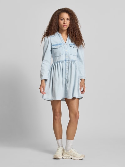 Frauenmodel trägt Mini-Jeanskleid aus reiner Baumwolle Modell 'CYRUS' von Only in heller Denim-Optik, Ausgestellt, Mini-Länge, V-Ausschnitt, Rückenpasse, Knopfleiste, Langarm mit schmaler Manschette, Ganzkörperansicht von vorne.
