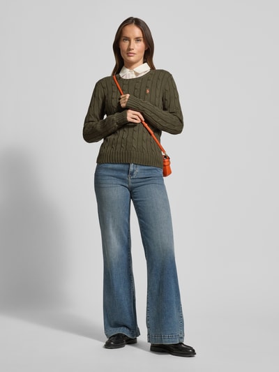 Frauenmodel trägt Strickpullover mit Zopfmuster Modell 'JULIANNA' von Polo Ralph Lauren, Reine Baumwolle, Slim Fit, Zopfmuster-Struktur, Label-Stitching, Rundhalsausschnitt, Olivgrün, Kombi mit blauer Jeans, Ganzkörperansicht von vorne.