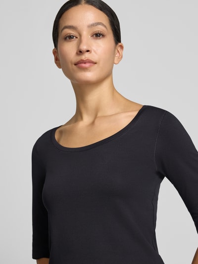 Modelka kobieca ma na sobie T-Shirt mit Ärmelaufschlag und Rundhalsausschnitt od Marc Cain, mieszanka bawełny z dodatkiem streczu, dopasowany krój, okrągły dekolt, zakładka na rękawach, detal z logo, długość tyłu w rozmiarze 36: 60 cm, zbliżenie górnej części ciała z przodu.