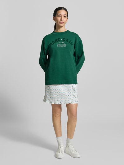 Frauenmodel trägt Relaxed Fit Sweatshirt mit Logo-Detail von Marc Cain in Dunkelgrün, Baumwoll-Mix, zu Rock, Ganzkörperansicht von vorne.