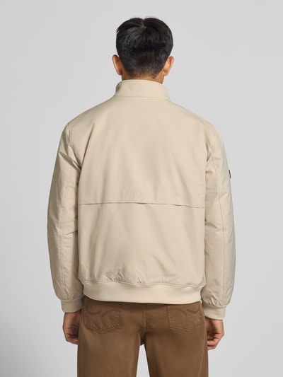 Männermodel trägt Regular Fit Jacke mit Logo-Stitching Modell 'HARRINGTON' von Superdry in Beige vor grauem Hintergrund, Ganzkörperansicht Rückansicht. Elastischer Material-Mix, Stehkragen, sichtbare Details am Rücken.