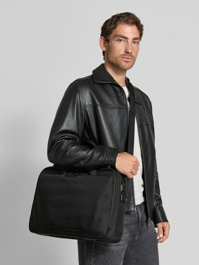 Męski model nosi czarną torbę na laptop Model 'Pro-DLX 6' od Samsonite z odpinanym paskiem na ramię, główną przegrodą na zamek błyskawiczny, krótkimi uchwytami, wewnętrznymi przegródkami i przednią przegródką, detal z logo; Szerokość w rozmiarze 1: 44 cm, Długość w rozmiarze 1: 31 cm, Głębokość w rozmiarze 1: 12 cm, Długość paska na ramię w rozmiarze 1: 128 cm; całość w odcieniach czerni, tekstura gładka, prostokątny kształt; widok całej sylwetki z przodu.