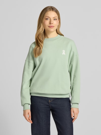 Modelka kobieca nosi Oversized Fit Sweatshirt aus reiner Bio-Baumwolle Modell 'ICONIC A ALIZAA' od Armedangels, miękka tekstura, kolor jednolity, okrągły dekolt, logo na piersi, długie rękawy z prążkowanym wykończeniem; widok całej sylwetki z przodu.