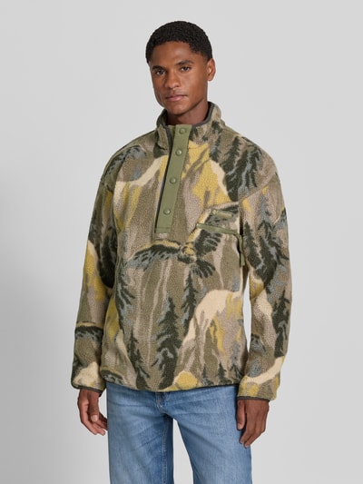 Männermodel trägt Fleecepullover mit Label-Patch Modell 'HELVETIA' von Columbia, prosty krój, stójka, naszywka z logo, kieszeń na piersi zapinana na zamek błyskawiczny, długość tyłu w rozmiarze M: 73 cm, długość rękawów w rozmiarze M: 60 cm, wzór kamuflażowy w odcieniach zieleni, żółci i beżu, tekstura miękka, Ganzkörperansicht von vorne.