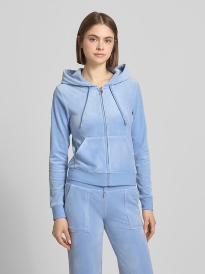 Frauenmodel trägt Slim Fit Sweatjacke aus Samt Modell 'Robertson' von Juicy Couture in Hellblau, mit Kapuze, Tunnelzug, Reißverschluss, Känguru-Tasche, Elastischer-Material-Mix, elastische Abschlüsse; Ganzkörperansicht von vorne.