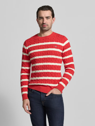 Männermodel trägt Slim Fit Pullover in Strick-Optik von Gant in Rot mit cremefarbenen Streifen, reine Baumwolle, Logo-Stitching, Ganzkörperansicht von vorne.