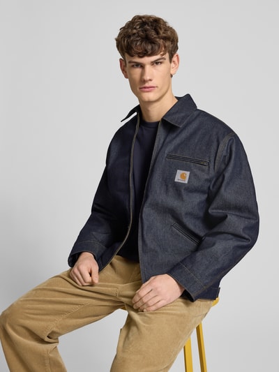 Männermodel trägt Jeansjacke mit Brusttasche und Label-Detail von Carhartt Work In Progress, dunkelblau, Kontrastnähte, Nahaufnahme Oberkörper. Regular Fit, Umlegekragen, Modell ‘DETROIT’, Reißverschluss, Eingrifftaschen, markantes Label, strukturierte Denim-Textur.