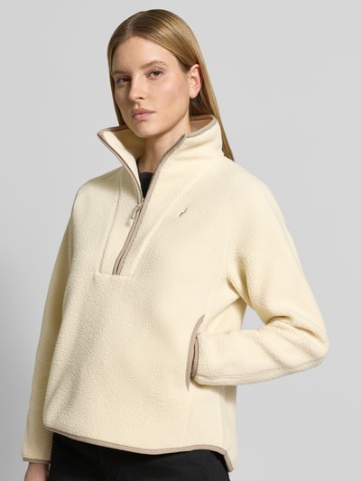 Modelka nosi Regular Fit Sweatshirt mit Stehkragen von PEAK PERFORMANCE, kremowa bluza, zamek błyskawiczny, wyhaftowane logo, krój regular fit, długość tyłu w rozmiarze S: 66 cm, szerokość ramion w rozmiarze S: 45 cm, szerokość klatki piersiowej w rozmiarze S: 57 cm, długość rękawów w rozmiarze S: 60 cm, tekstura miękka, gładka, z przodu, zbliżenie na górną część ciała.