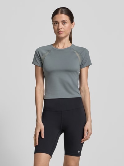 Frauenmodel trägt T-Shirt mit Mesh-Einsätzen und Logo von Under Armour in Grau, Cropped, Material-Mix mit Stretch-Anteil, Rundhalsausschnitt, Rückenlänge bei Größe S 45 cm, Ganzkörperansicht von vorne.