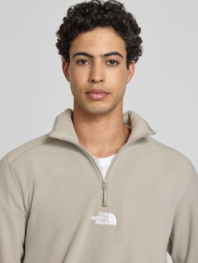 Męski model nosi Fleecepullover mit Reißverschluss Modell 'GLACIER' von The North Face, krój regular fit, stójka, wyhaftowane logo, zamek błyskawiczny, beżowy, tekstura polaru, długość tyłu w rozmiarze M: 70 cm, długość rękawów w rozmiarze M: 67 cm, Nahaufnahme górnej części ciała.