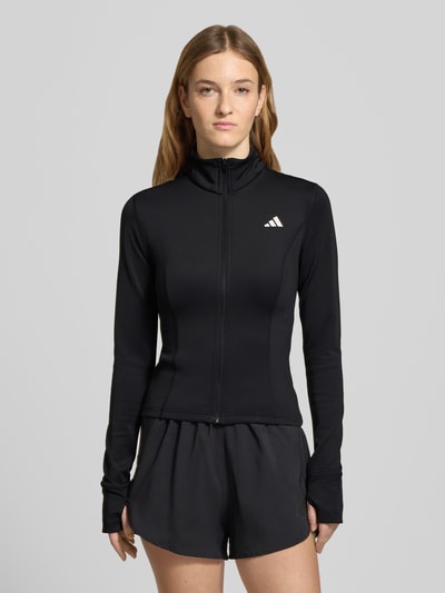 Frauenmodel trägt Slim Fit Jacke mit Stehkragen von adidas Sportswear in Schwarz, elastisches Material, Logo-Print, Ganzkörperansicht von vorne.