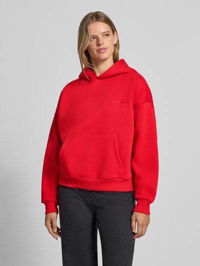Frauenmodel trägt Kapuzenpullover aus Baumwoll-Mix von REVIEW in leuchtendem Rot mit Känguru-Tasche, lockere Passform, Ganzkörperansicht von vorne.