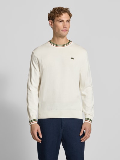 Männermodel trägt Regular Fit Pullover aus reiner Wolle von Lacoste in Creme mit grünen und roten Streifen, Ganzkörperansicht von vorne. Herren Pullover von Lacoste