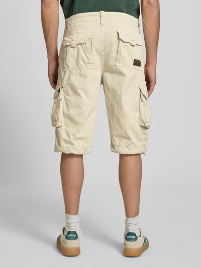 Mężczyzna nosi Bermudas mit Pattentaschen und Logo-Stitching od Alpha Industries, beżowy kolor, bawełna, krój straight fit, widok całej sylwetki z tyłu. Szerokość w rozmiarze 32: 44 cm, długość nogawki od wewnątrz w rozmiarze 32: 33 cm, kieszenie boczne i na pośladkach, kieszenie z klapką na rzep, logo-stitching