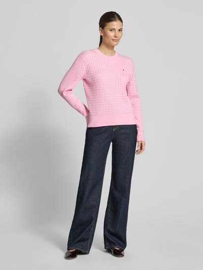 Frauenmodel trägt Slim Fit Pullover aus reiner Baumwolle von Tommy Hilfiger in Hellrosa, Zopfmuster, Crew Neck, lange Ärmel, unifarben, Strickoptik, Ganzkörperansicht von vorne.