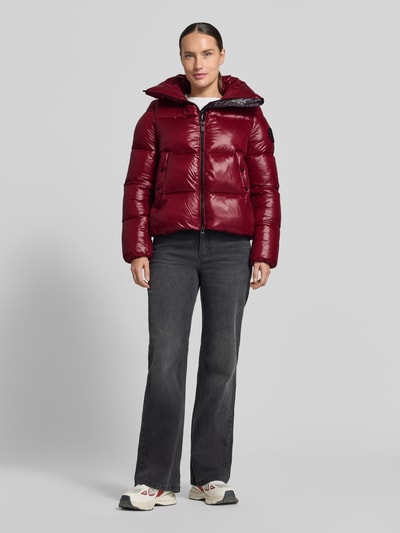 Frauenmodel trägt glänzende, bordeauxrote Regular Fit Steppjacke mit Stehkragen, Modell 'Isla' von Save The Duck, Ganzkörperansicht von vorne. Damen Steppjacke, Stepp-Optik, Zweiwege-Reißverschluss, Eingrifftaschen, Rückenlänge 56 cm, Schulterbreite 14 cm, Brustweite 51 cm, Ärmellänge 66 cm.